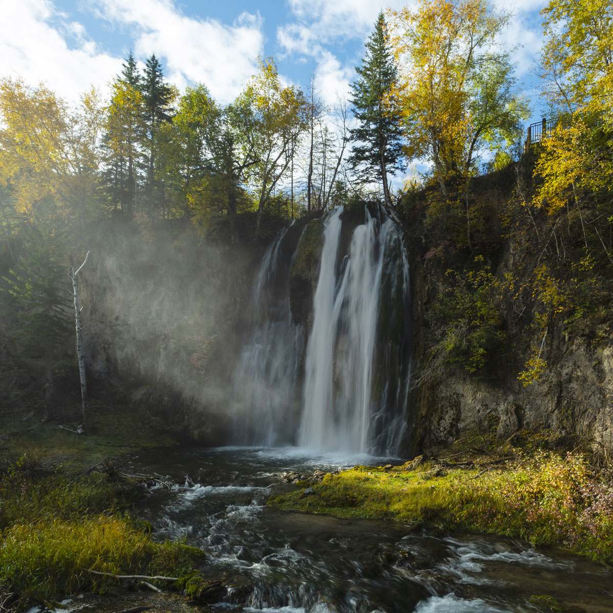White water crashes down Spearfish Falls casting a silvery mist in the golden early morning light amid trees of yellow and green Autumn colors.